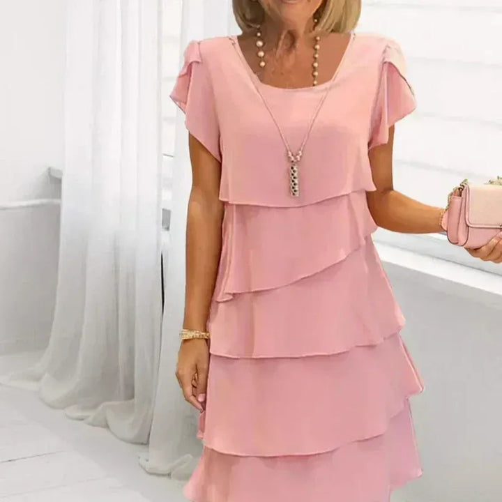 Close up front view of a woman wearing a pink ruffled wavy cut dress with a layered design, holding a handbag