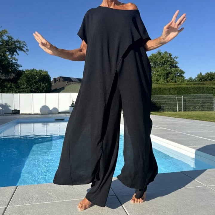 Model wearing a black asymmetrical off shoulder top and wide leg pants set by a pool, arms outstretched