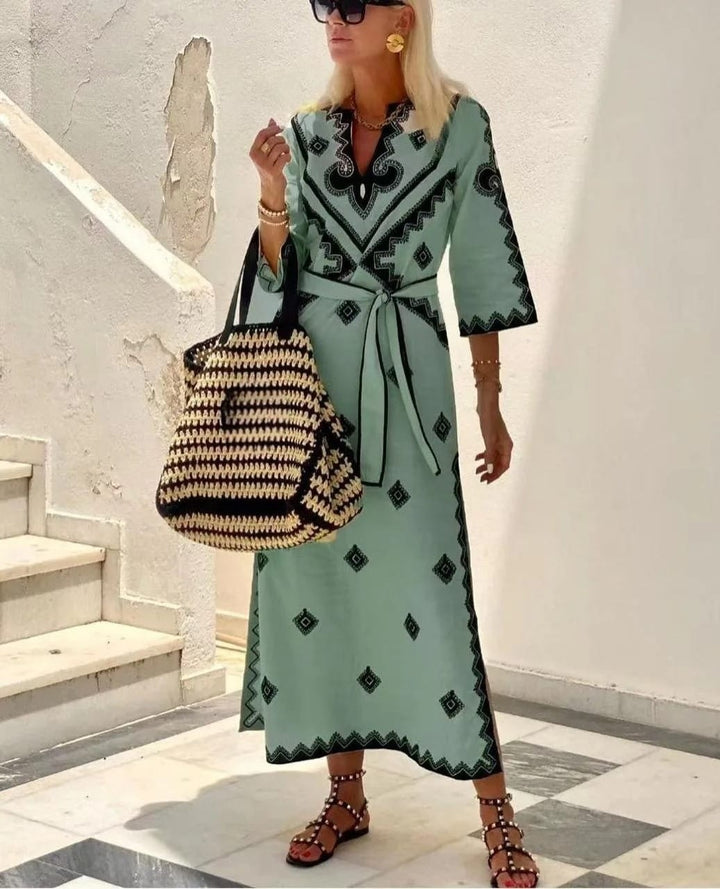 Side view of a model wearing a printed waist tie midi dress with a woven bag and black studded sandals
