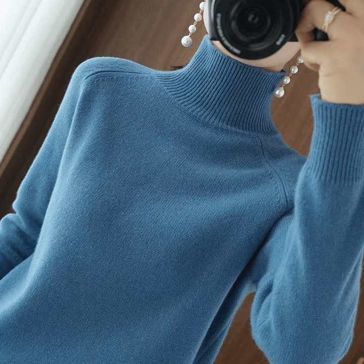Close up of a model wearing the teal turtleneck jumper, holding a camera and long pearl earrings