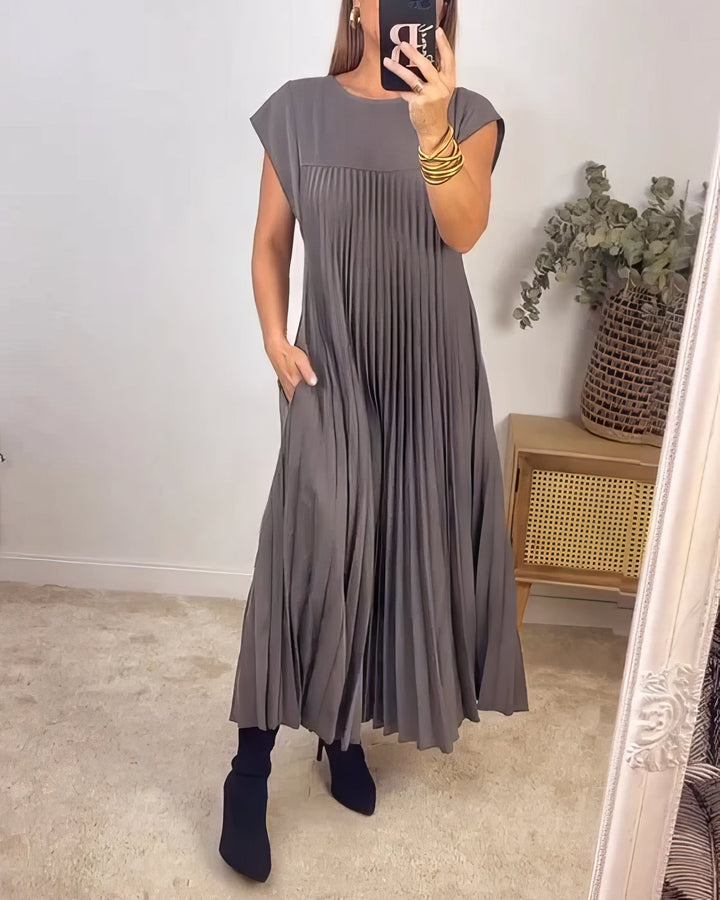 Sleeveless pleated dress, front view worn by model, hand in pocket, holding phone, gold cuffs, earrings and black boots