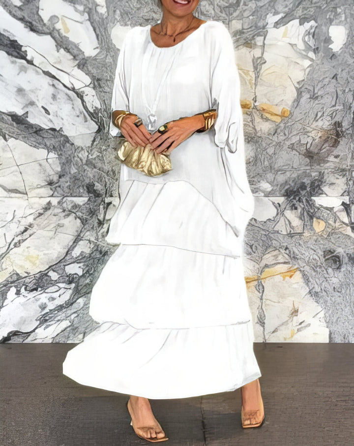 Front view of a woman wearing a white Artistic Flow Dress with tiered skirt, gold clutch and clear heels