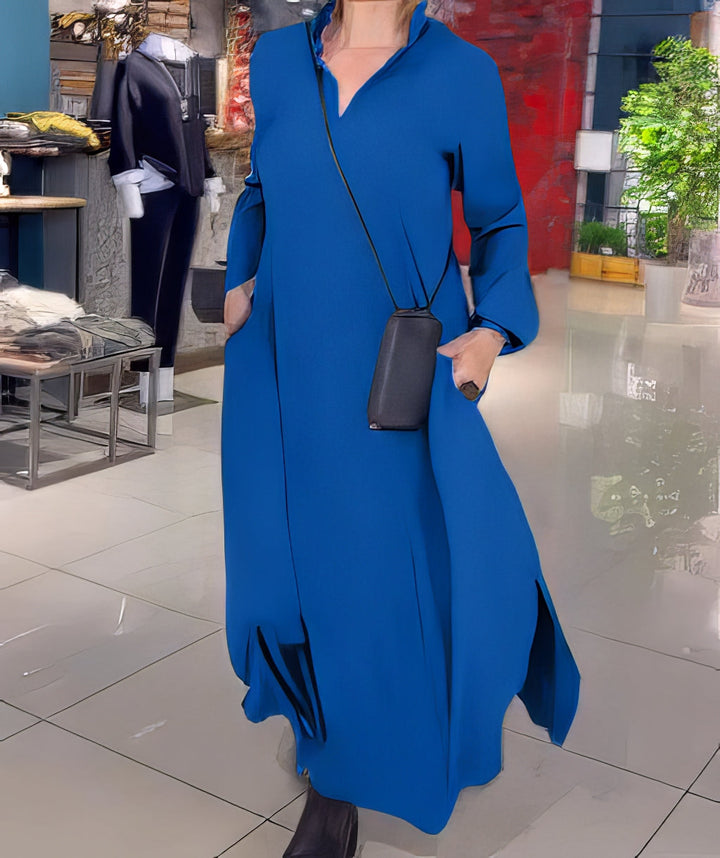 Front view, model in a blue stretch maxi dress with side slits, hands in pockets carrying a small phone bag