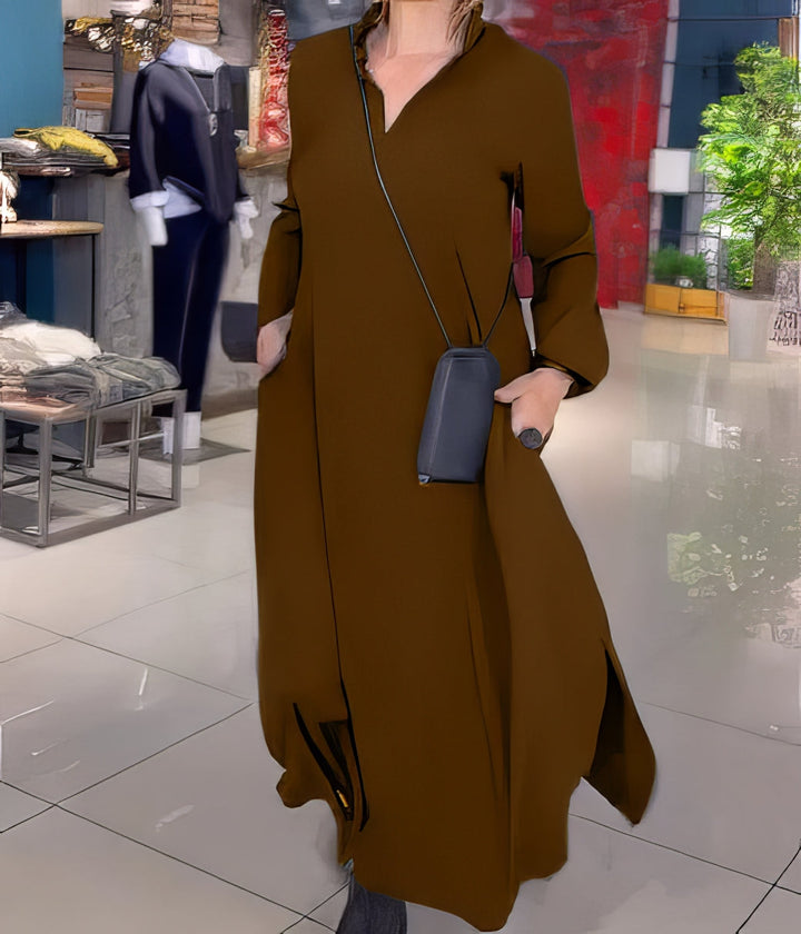 Three quarter view, model in a brown stretch maxi dress with side slits, hands in pockets carrying a small bag