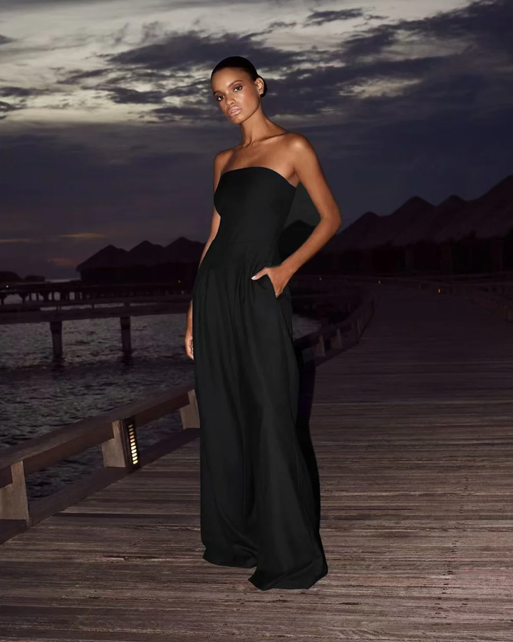 Front view of a model wearing a strapless black Luxe Flow Jumpsuit on a wooden pier at dusk