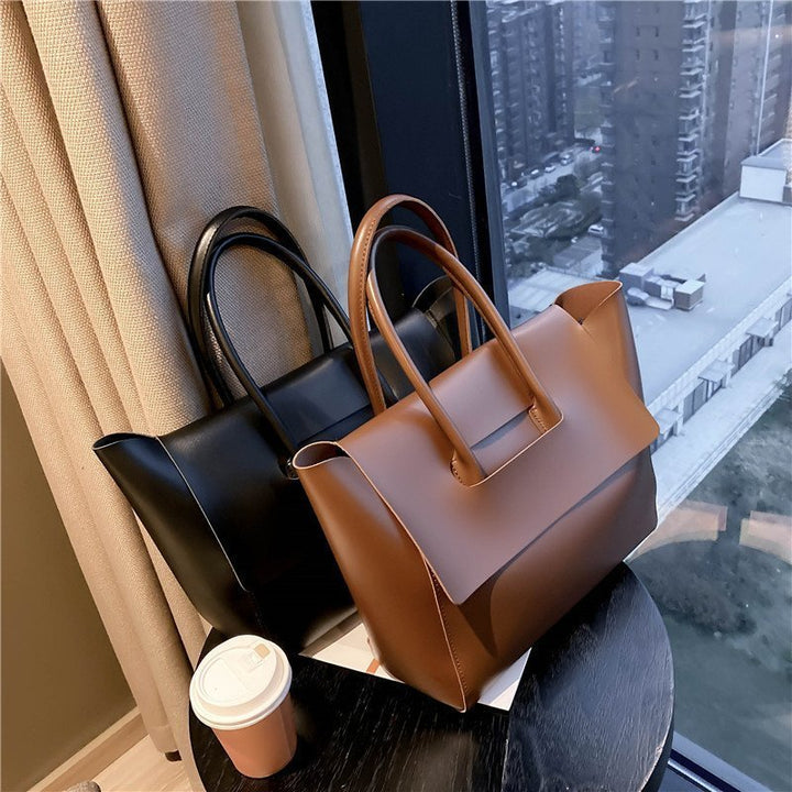 Front view of two Geneva vegan leather handbags, one black and one tan, on a table by a window