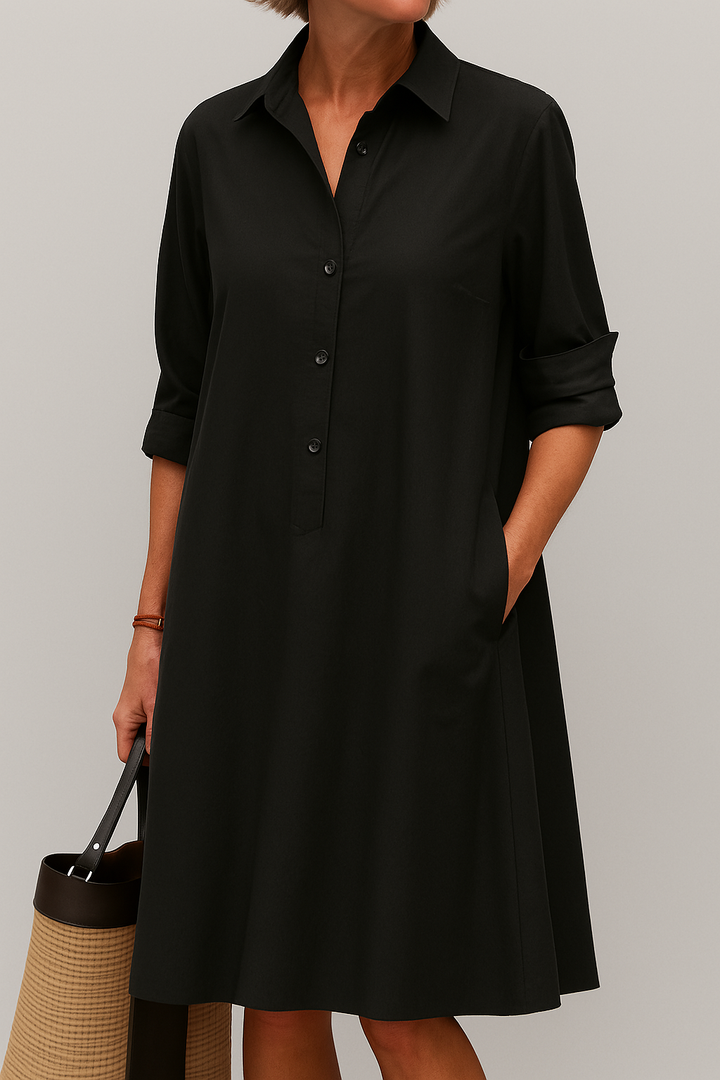 Front view, model wearing a black button-down shirt dress with rolled sleeves, one hand in a pocket, holding a straw bag