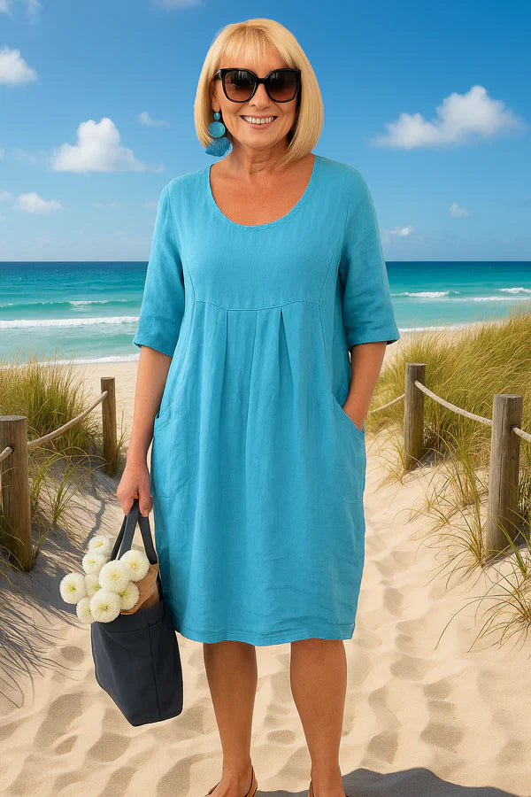 Front view of a smiling woman in a blue linen summer dress with pockets, holding a bag of flowers on a beach