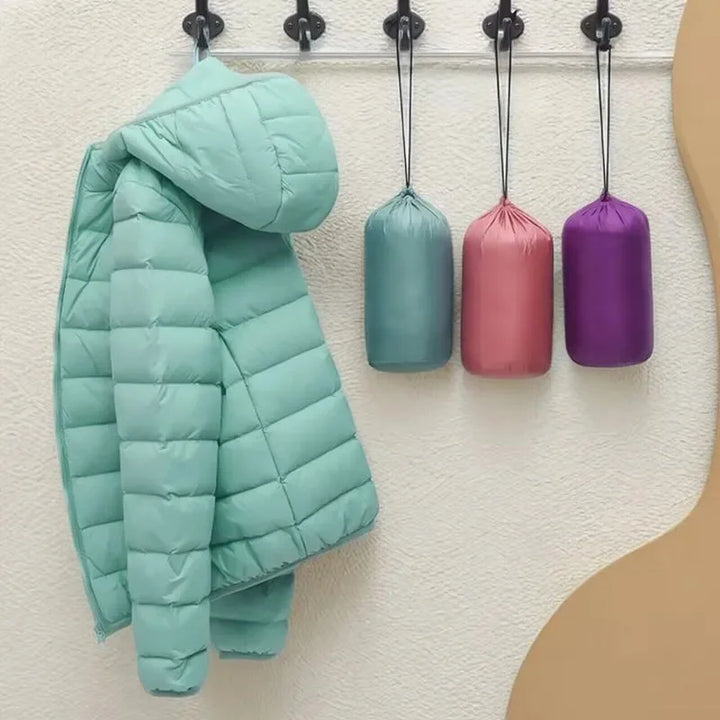 Front view of a mint hooded lightweight puffer jacket on a hook, with three rolled jackets in bags