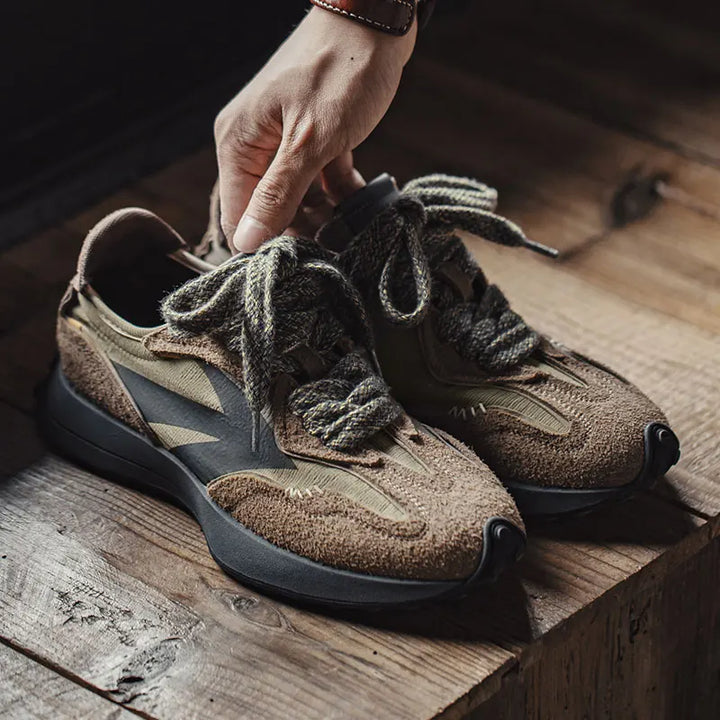 Front view of Noah 70s retro suede sneakers in brown and olive with a hand holding one sneaker