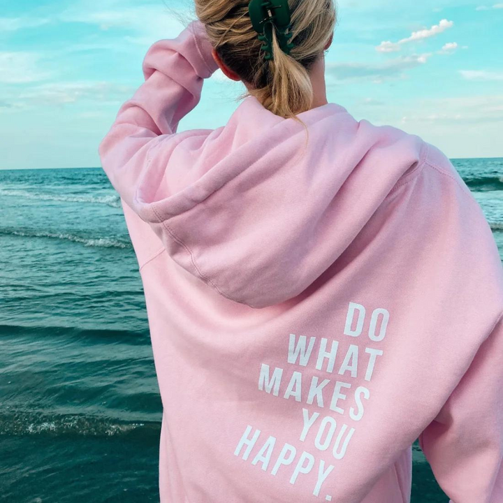 Back view, model wearing a pink relaxed knit pullover hoodie with "DO WHAT MAKES YOU HAPPY" text, by the ocean