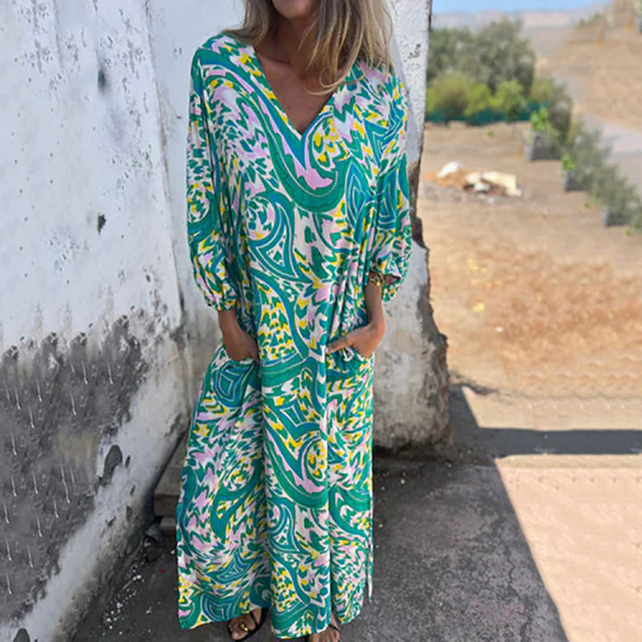 Front view, model wearing a V-neck maxi dress in a green, yellow and pink abstract print with hands in pockets