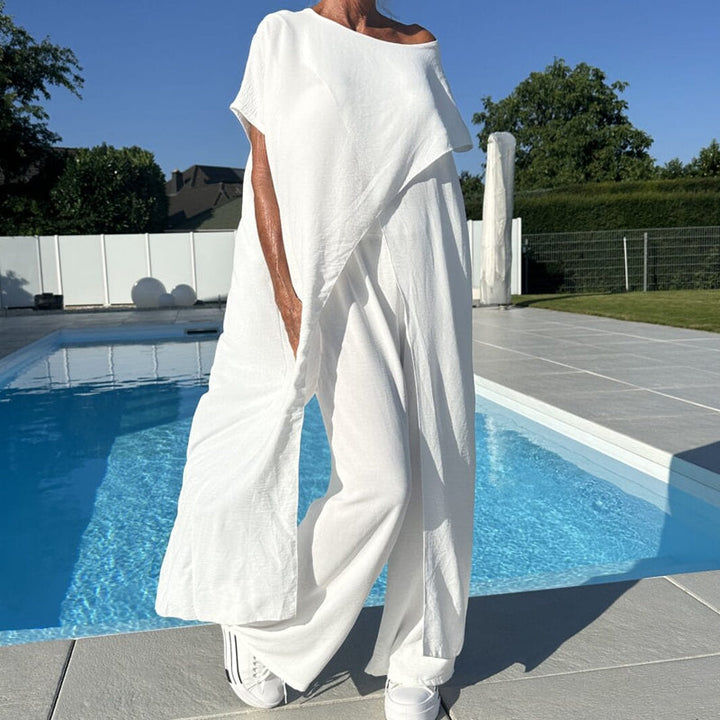 Front view of a model wearing a white asymmetrical off shoulder top and wide leg pants set by a pool
