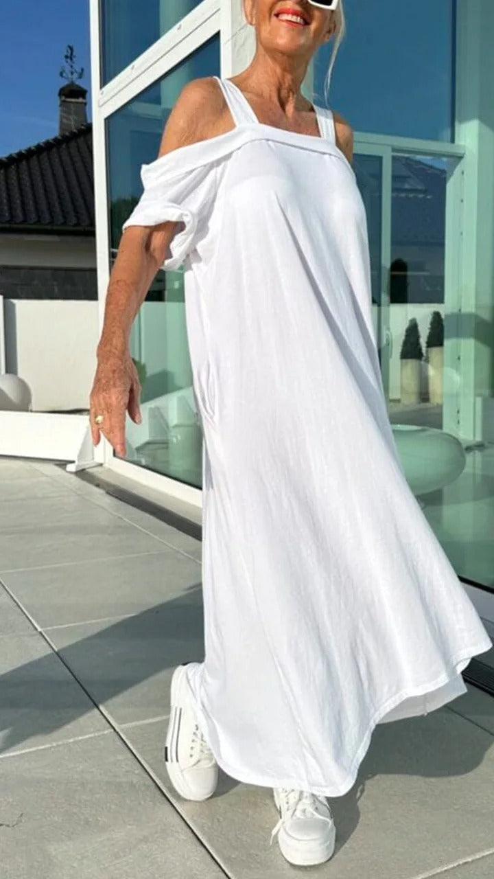 Front view, model wearing a white Brianna maxi dress with open shoulders and white sneakers on a balcony